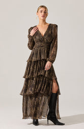 A woman is wearing a long, flowy dress with a leopard print pattern. The dress has puffed long sleeves, a V-neckline, and a fitted waist. It features multiple tiered ruffles down the length, with a high slit on one side, revealing knee-high black boots with high heels.
